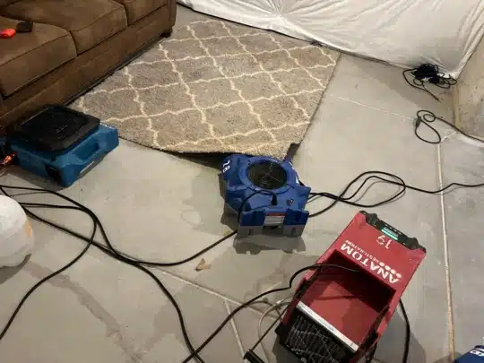 Multiple air movers and drying equipment are arranged on a basement concrete floor near a partially lifted rug and a brown sofa.
