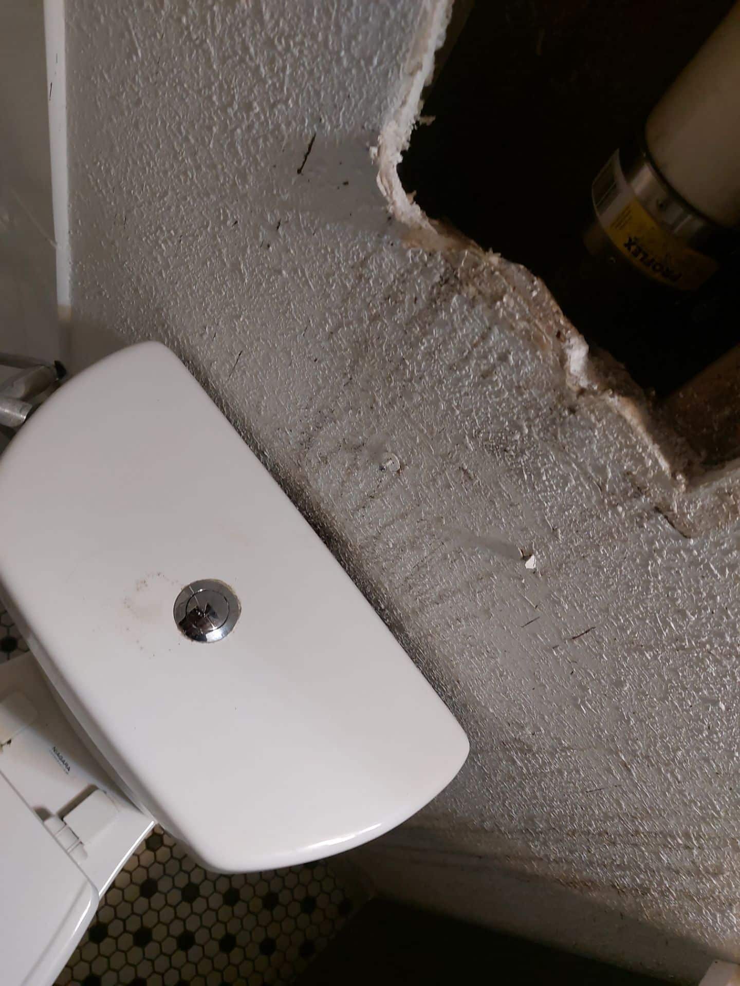 A damaged bathroom wall with a hole exposing plumbing behind a toilet tank