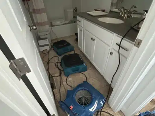 Three blue air movers and drying equipment are placed on the floor of a residential bathroom to dry water damage, with visible sink, cabinets, and toilet in the background