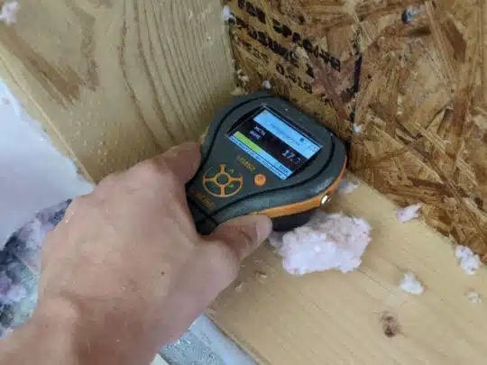 A hand holds a moisture meter against an exposed wooden wall with insulation nearby.