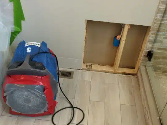 A blue industrial air mover is placed on a bathroom floor next to an opened wall cavity with exposed studs and electrical wiring.