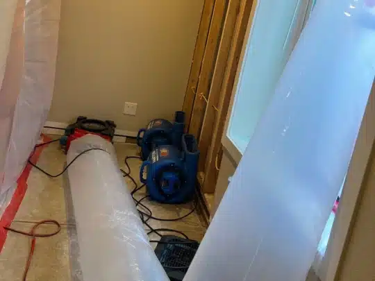 Two blue industrial air movers and drying equipment are set up in a residential room with exposed wall studs and plastic sheeting for water damage restoration.