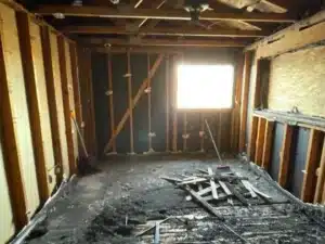 A room stripped down to its framing shows significant fire damage. The floor is covered in black debris, and a shovel and broom are visible.