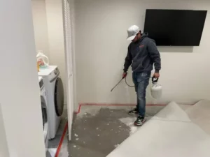 A restoration professional spraying a solution on a concrete floor in a laundry room with a washer and television.