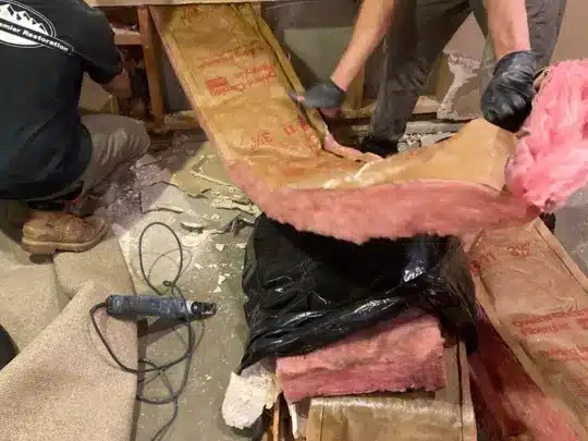 Two technicians are removing damaged insulation from a wall in a room, with tools and debris scattered around