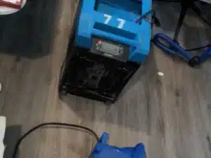A blue dehumidifier and a blue industrial fan on a wooden floor in a room with a desk, chair, and blanket in the background