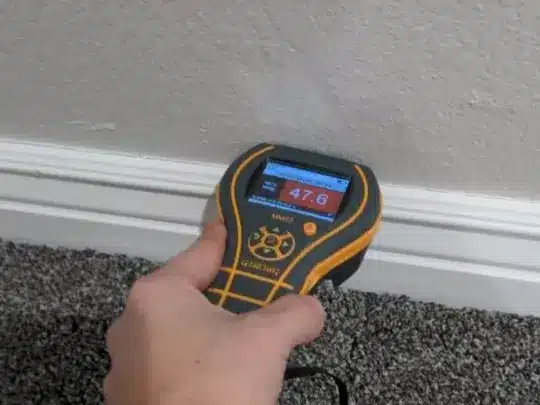 A person holding a moisture detection device against a wall near the baseboard, displaying a reading of 47.6