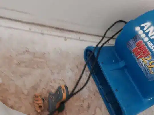 A blue air mover with black cables is placed on a wooden floor near a white wall.