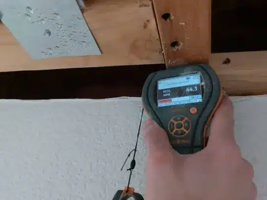 A person holding a moisture meter displaying "44.3" against a wooden beam