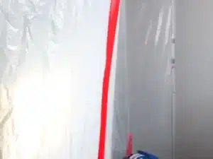 A doorway is sealed with a transparent plastic ZipWall kit, secured with red tape. A blue dehumidifier is visible near the bottom, and a gray toolbox is in the foreground