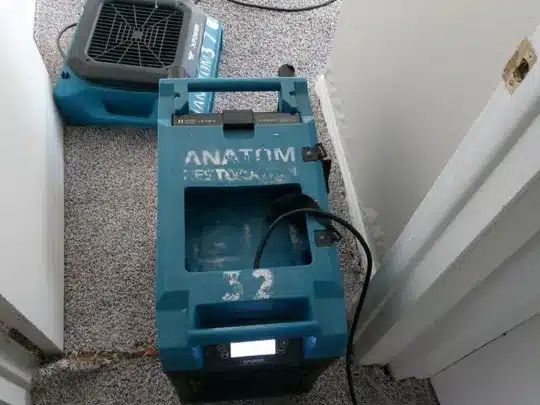 Two blue drying machines on a carpeted floor, connected by cables, positioned near a door in a room