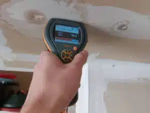 A person holding a moisture meter against a water-stained ceiling in an indoor setting