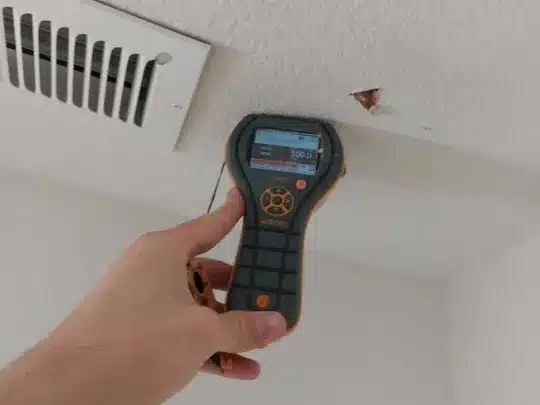 A hand holding a Protimeter MMS3 device against a damaged ceiling