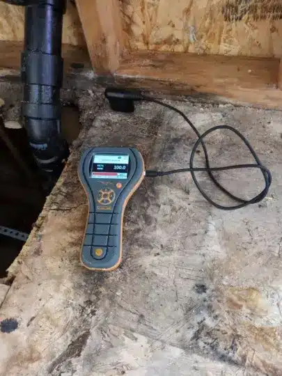 Moisture meter showing 100 on damaged wooden floor.