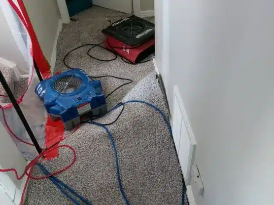 Restoration technicians' equipment, including air movers and hoses, is placed on a carpeted hallway near a door