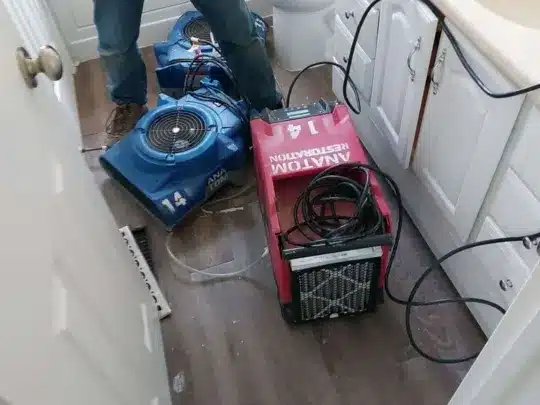 A person standing in a bathroom, with a blue and a red drying machine on the floor, connected by cables, near a sink and toilet