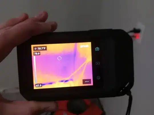 A person holding a thermal imaging camera, pointing it at a wall, with the screen showing a heat map and temperature readings