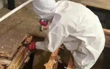 Photo of a worker in a white protective suit with a mask, working on a bare wooden floor during a renovation, holding a tool in his hand