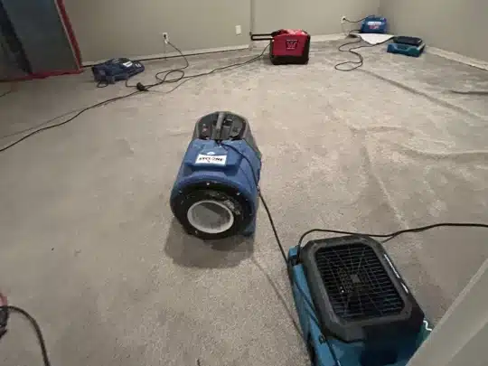 Multiple industrial air movers and drying equipment are placed on a carpeted floor in an empty room during a water damage restoration process.