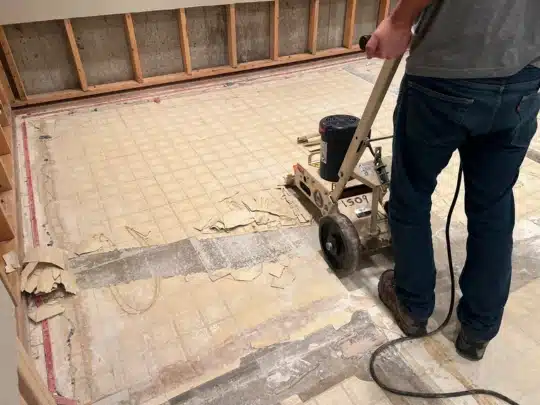 A restoration technician operates a floor scraper machine to remove old vinyl flooring in a room with exposed wall framing and visible water damage.