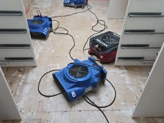 A set of four air movers and a red dehumidifier placed on a stripped wooden subfloor between white cabinet frames