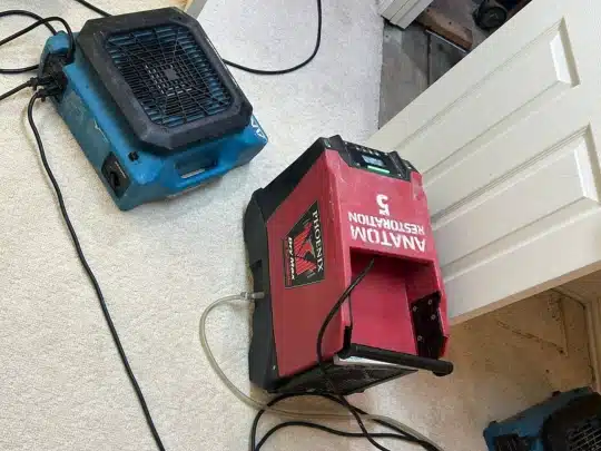 Restoration equipment including a red dehumidifier labeled Anatom Restoration and a blue air mover on wet carpet