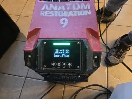 Close-up of a red ANATOM RESTORATION dehumidifier displaying humidity and temperature readings on a digital screen