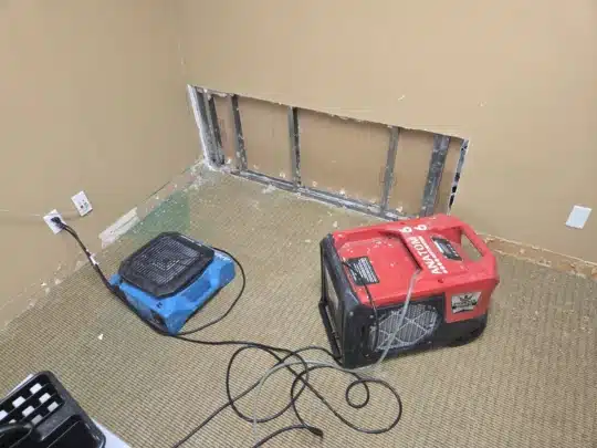 Two drying machines placed near a wall with the lower section of drywall cut out, exposing metal framing