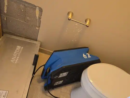 A commercial restroom with a toilet, a removed section of wall exposing concrete blocks, a metal drying panel labeled "Anatom," and a blue air mover marked "62."