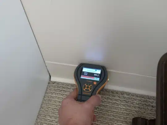 A technician holds a moisture meter against the base of a wall, showing a reading of 99.1 on the screen