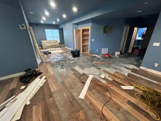 Partially removed flooring in a living room with tools, wires, and wood planks scattered during a renovation project.