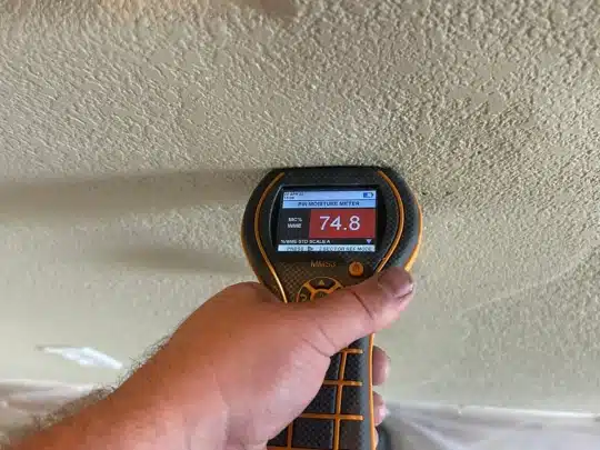 A technician holding a moisture meter against a textured ceiling, showing a high reading of 74.8 percent