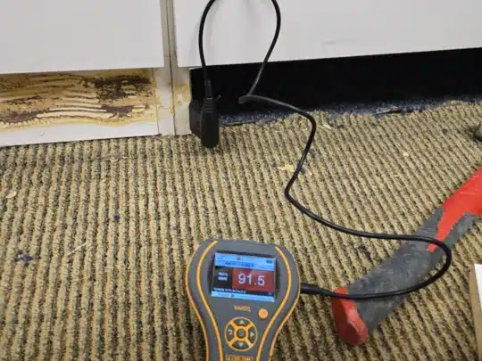 Moisture meter showing high reading on wet carpet near a damaged cabinet base