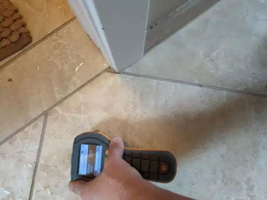 A technician measures moisture levels on a tiled floor near a bathroom door using a handheld meter