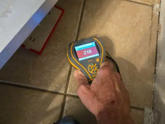 Technician holding a handheld moisture meter on a tiled floor, with the screen showing a reading of 219
