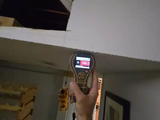 A technician holding a moisture meter against a ceiling, showing a reading of 100 in a residential interior