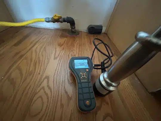 A moisture meter placed on hardwood flooring next to a gas line and a kitchen sink pipe