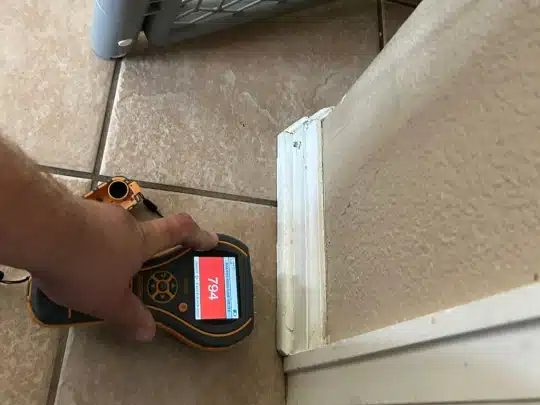 Technician holding a handheld moisture meter against a wall and tile floor