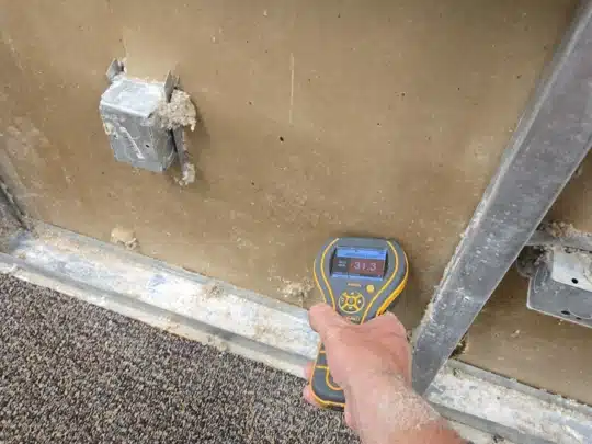 Hand holding a moisture meter against a wall with visible electrical outlets and damaged drywall