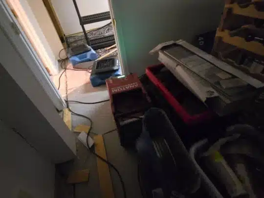 A cluttered storage area with bins, tools, and equipment, with air movers set up in an adjacent hallway