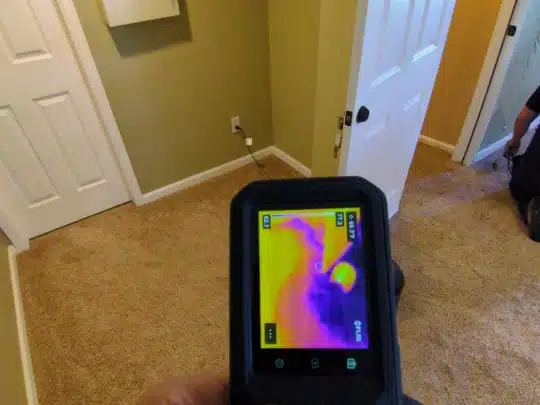 A technician holding a thermal imaging camera showing heat patterns on a wall inside a carpeted room