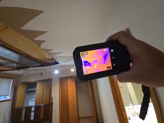 A technician holding a thermal imaging camera pointed at a ceiling with visible damage and exposed beams