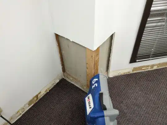 Drywall cut out near the floor with exposed framing and a drying machine placed in the corner of the room
